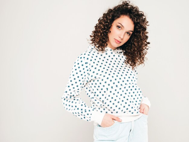 Retrato de hermosa modelo sonriente con peinado afro rizos vestido con ropa hipster de verano. Chica despreocupada sexy posando en estudio sobre fondo gris. Mujer divertida y positiva de moda