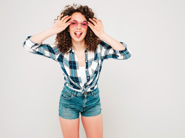 Retrato de hermosa modelo sonriente con peinado afro rizos vestido con ropa hipster de verano. Chica despreocupada sexy posando en estudio sobre fondo gris. Mujer de moda muestra lengua