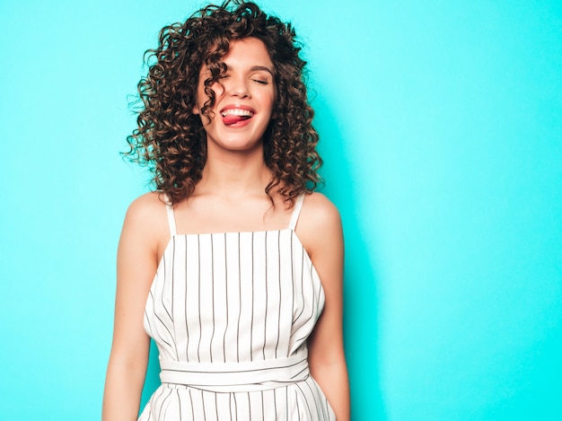 Retrato de hermosa modelo sonriente con peinado afro rizos vestido con ropa hipster de verano. Chica despreocupada sexy posando junto a la pared azul. Mujer divertida y positiva de moda. Muestra lengua