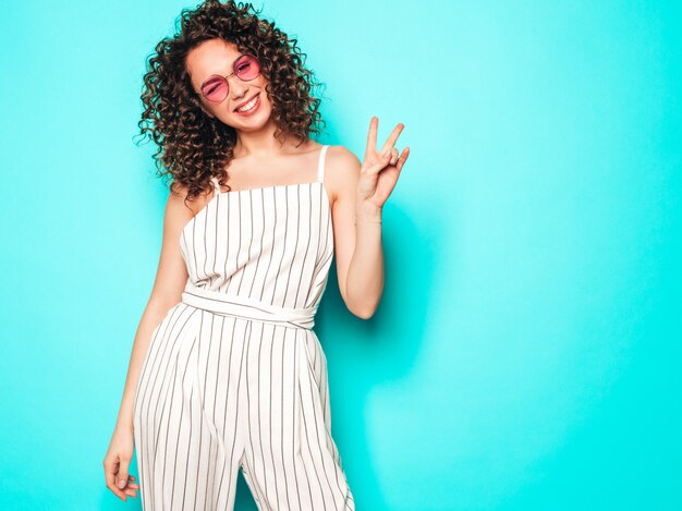 Retrato de hermosa modelo sonriente con peinado afro rizos vestido con ropa hipster de verano. Chica despreocupada sexy posando junto a la pared azul. Mujer divertida y positiva de moda