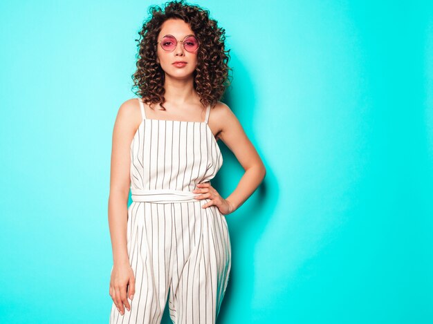 Retrato de hermosa modelo sonriente con peinado afro rizos vestido con ropa hipster de verano. Chica despreocupada sexy posando junto a la pared azul. Mujer divertida y positiva de moda