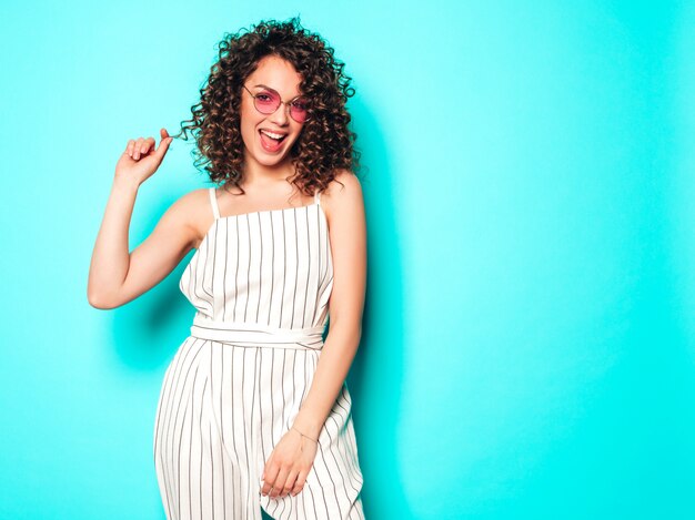 Retrato de hermosa modelo sonriente con peinado afro rizos vestido con ropa hipster de verano. Chica despreocupada sexy posando junto a la pared azul. Mujer divertida y positiva de moda