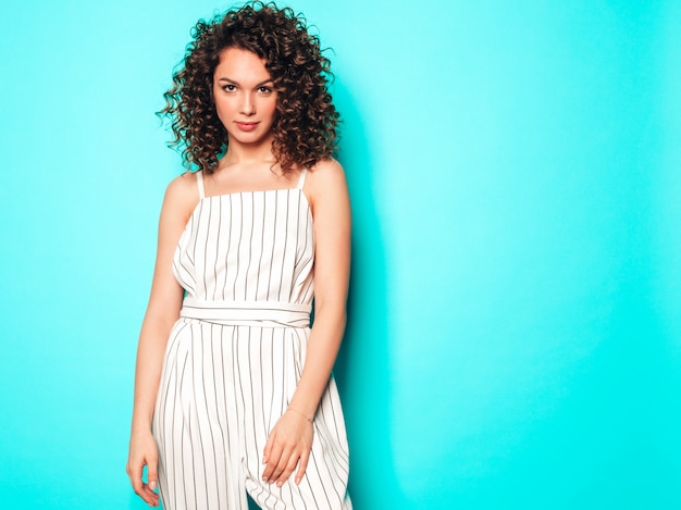 Retrato de hermosa modelo sonriente con peinado afro rizos vestido con ropa hipster de verano. Chica despreocupada sexy posando junto a la pared azul. Mujer divertida y positiva de moda