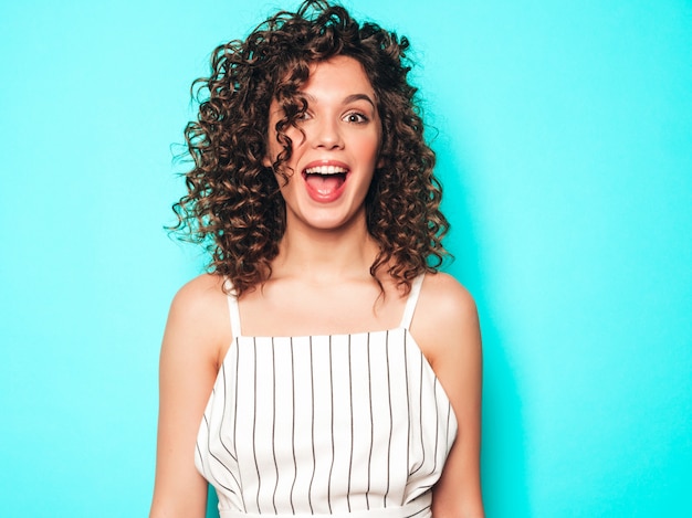 Retrato de hermosa modelo sonriente con peinado afro rizos vestido con ropa hipster de verano. Chica despreocupada sexy posando junto a la pared azul. Mujer divertida y positiva de moda