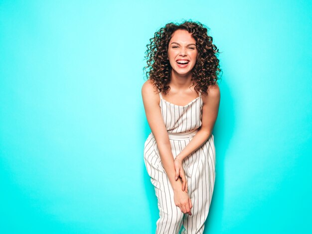 Retrato de hermosa modelo sonriente con peinado afro rizos vestido con ropa hipster de verano. Chica despreocupada sexy posando junto a la pared azul. Mujer divertida y positiva de moda