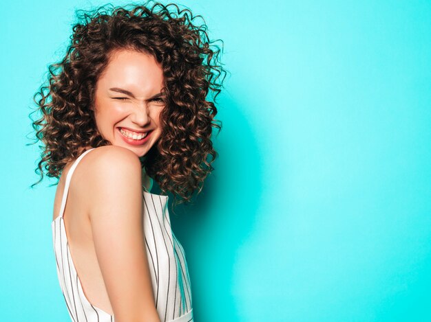 Retrato de hermosa modelo sonriente con peinado afro rizos vestido con ropa hipster de verano. Chica despreocupada sexy posando junto a la pared azul. Mujer divertida y positiva de moda