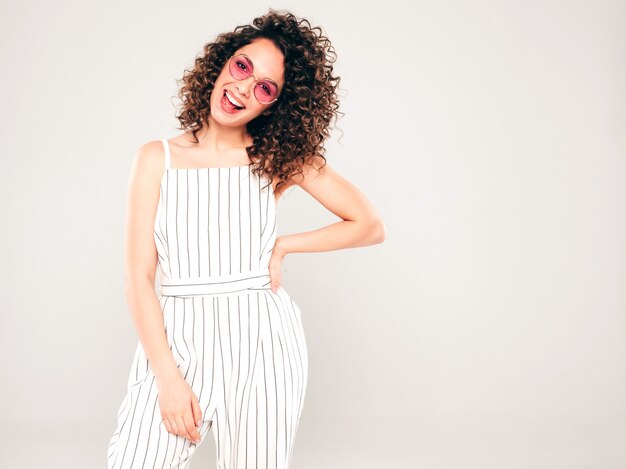 Retrato de hermosa modelo sonriente con peinado afro rizos vestido con ropa hipster de verano. Mujer divertida y positiva de moda en gafas de sol