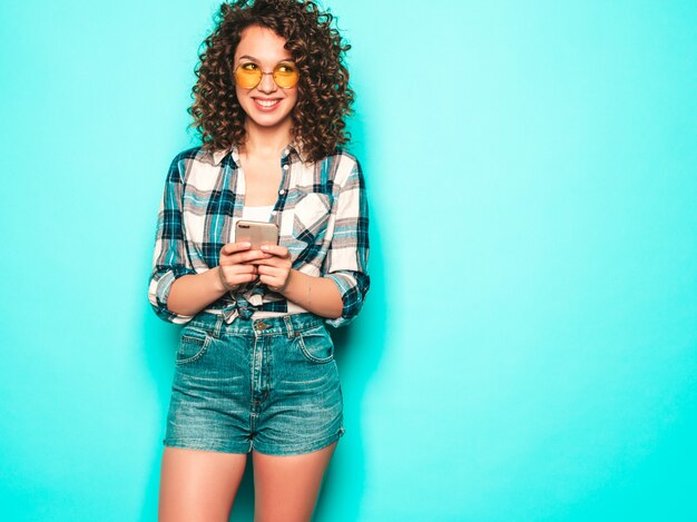 Retrato de hermosa modelo sonriente con peinado afro rizos vestido con ropa de verano. Chica despreocupada posando cerca de la pared azul. La mujer usa su teléfono móvil y escribiendo sms. Ella busca ventas en la tienda