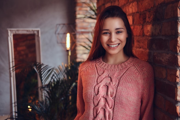 Retrato de una hermosa morena encantadora vestida con ropa elegante sonriendo mientras se apoya contra una pared de ladrillos en una habitación con interior de loft.