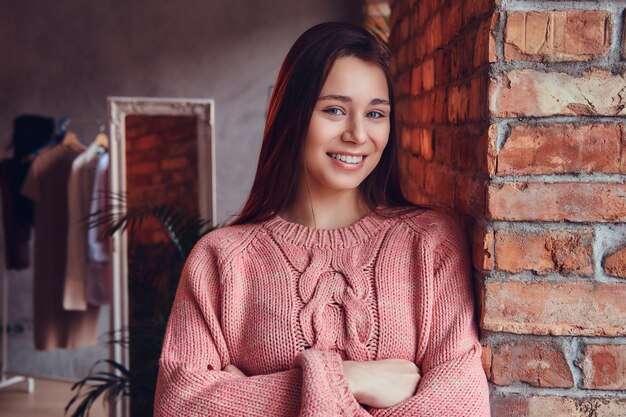 Retrato de una hermosa morena encantadora vestida con ropa elegante sonriendo mientras se apoya contra una pared de ladrillos en una habitación con interior de loft.