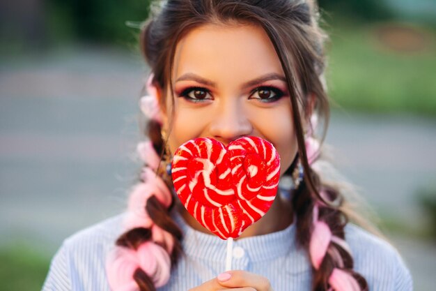 Retrato de una hermosa morena mirando a la cámara, escondiendo la cara con un corazón rojo caramelo en un palo. Mujer bonita y elegante con maquillaje y coloridas trenzas rosas. Estado de ánimo juguetón romántico.