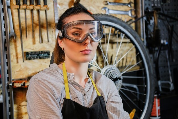 Retrato de una hermosa morena con ropa de trabajo, delantal y gafas, parada en un taller contra herramientas de pared.