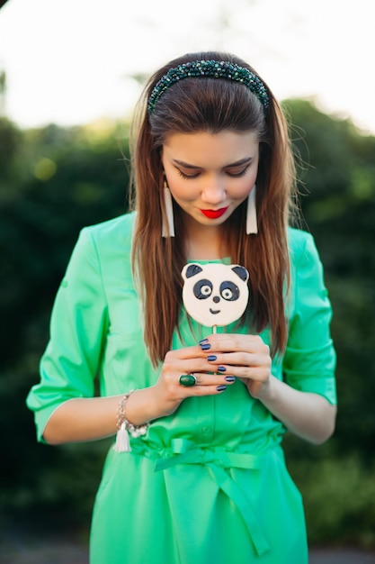 Retrato de una hermosa morena con vestido verde, comiendo dulces como panda en un palo y mirando al costado. Mujer bonita y elegante con labios rojos, aretes largos y banda para el cabello en el parque. Hora de verano.