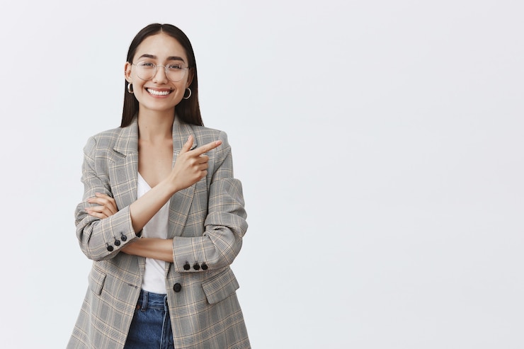 Foto gratuito retrato de hermosa mujer adulta despreocupada con gafas y chaqueta sobre camiseta blanca, apuntando a la esquina superior derecha y sonriendo con dientes brillantes