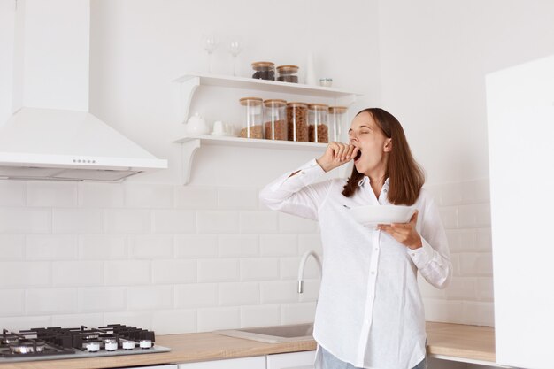 Retrato de hermosa mujer adulta joven de pelo oscuro con sueño desayunando en la cocina, de pie con el brazo levantado, bostezando, tapándose la boca con la mano.