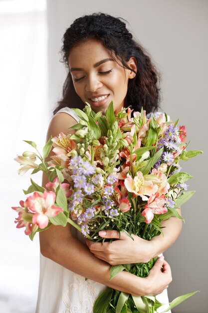 Retrato de hermosa mujer africana tierna en vestido blanco sonriendo con ramo de flores. Ojos cerrados.