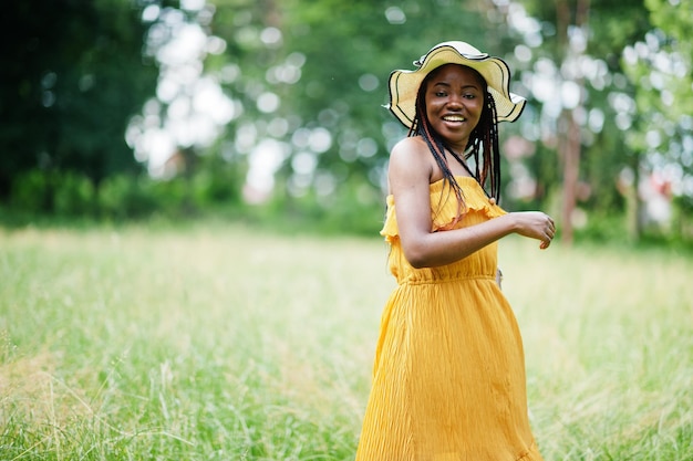 Retrato de hermosa mujer afroamericana de 20 años vestida con vestido amarillo y sombrero de verano posando en la hierba verde en el parque