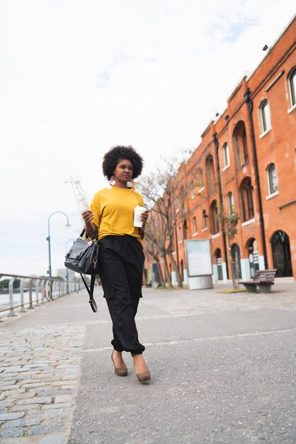 Retrato de hermosa mujer afroamericana caminando y sosteniendo una taza de café al aire libre en la calle. Concepto urbano.