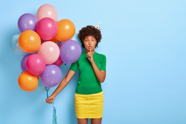Retrato de hermosa mujer afroamericana hace gesto de silencio, sostiene un montón de globos de colores, viste camiseta verde y falda amarilla, dice secreto