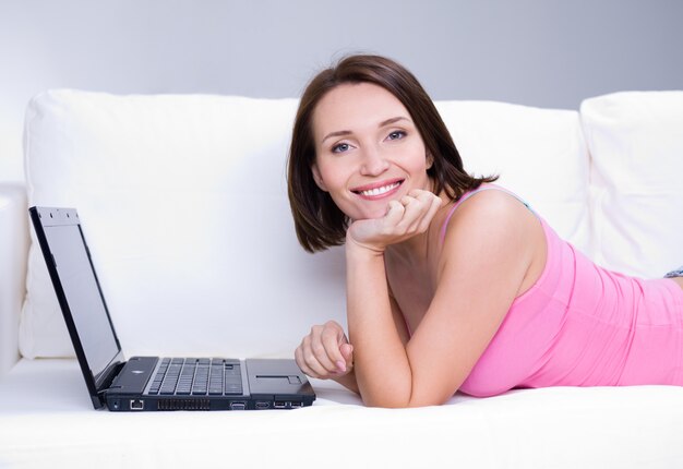 Retrato de hermosa mujer alegre con sonrisa feliz tumbado en el sofá con el portátil