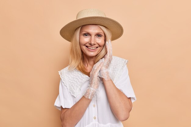 Retrato de hermosa mujer aristocrática con apariencia agradable sonríe felizmente mantiene las manos juntas viste un vestido blanco fedora y guantes de encaje expresa poses de emociones positivas contra la pared beige