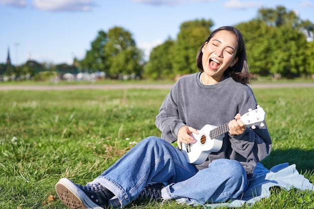 Retrato de una hermosa mujer asiática cantando tocando la guitarra ukelele en el parque sentada sola en el césped en s