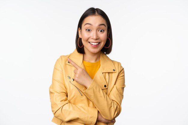 Retrato de una hermosa mujer asiática expresa interés y entusiasmo por el descuento de la tienda de publicidad señalando con el dedo a la izquierda mirando asombrado de pie sobre fondo blanco