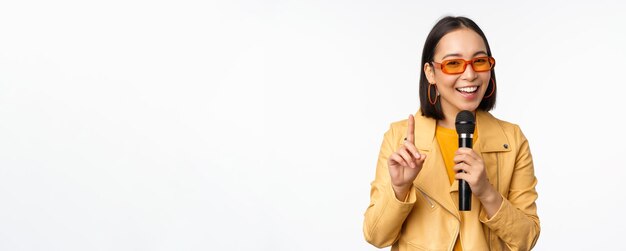 Retrato de una hermosa mujer asiática con gafas de sol, una chica elegante cantando dando un discurso con un micrófono sosteniendo un micrófono y sonriendo de pie sobre fondo blanco