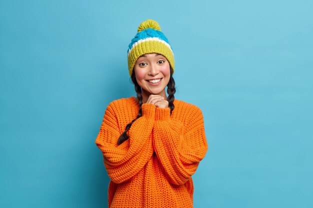 Retrato de hermosa mujer asiática con gran sonrisa mantiene las manos juntas estando de buen humor viste gorro de punto de invierno y suéter naranja posa contra la pared azul