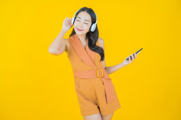 Retrato de hermosa mujer asiática joven con auriculares y teléfono inteligente para escuchar música sobre fondo amarillo