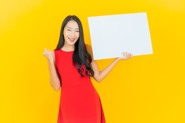 Retrato hermosa mujer asiática joven sonrisa con cartelera blanca vacía en la pared amarilla