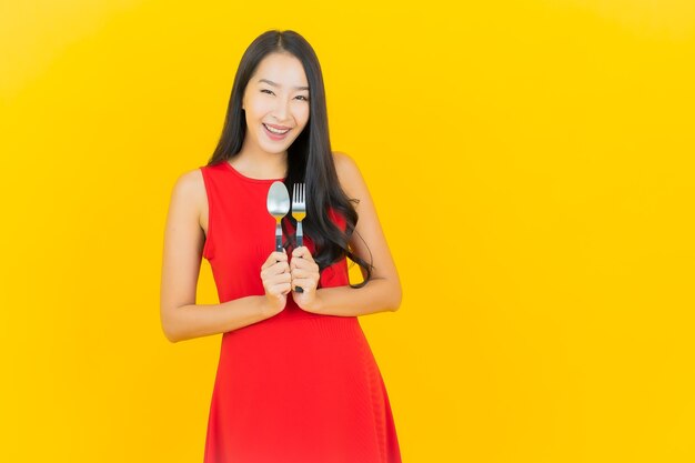 Retrato hermosa mujer asiática joven sonrisa con cuchara y tenedor en la pared amarilla