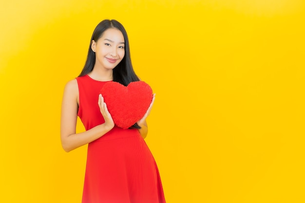 Retrato hermosa mujer asiática joven sonrisa con forma de almohada de corazón en la pared amarilla