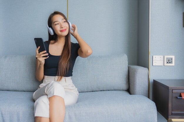 Retrato hermosa mujer asiática joven usar teléfono móvil inteligente con auriculares para escuchar música en el sofá en la sala de estar