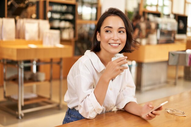 Foto gratuita retrato de una hermosa mujer asiática con un teléfono inteligente relajándose en un café sentado y disfrutando de un café mientras usa el teléfono móvil