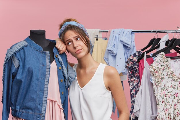 Retrato de hermosa mujer cansada apoyándose en el maniquí mientras está de pie en boutigue, agotado probándose ropa, teniendo dudas sobre qué comprar. Infeliz mujer compradora en tienda de ropa siente aburrimiento