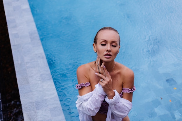 Retrato de hermosa mujer caucásica en bikini y camisa blanca en piscina azul bajo la lluvia