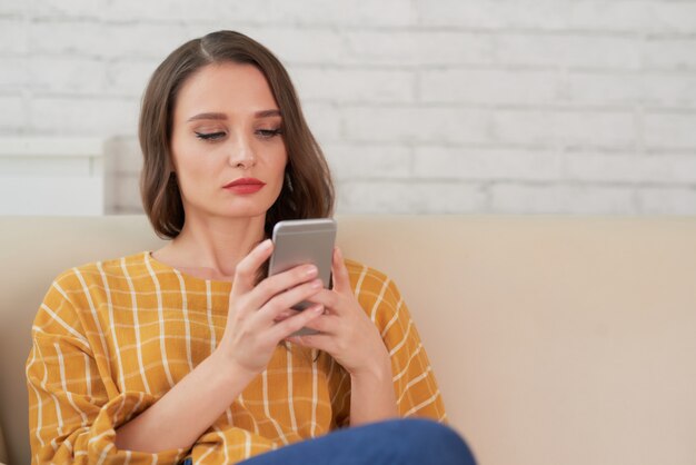 Retrato de hermosa mujer caucásica enviando mensajes de texto por teléfono sentado en el sofá