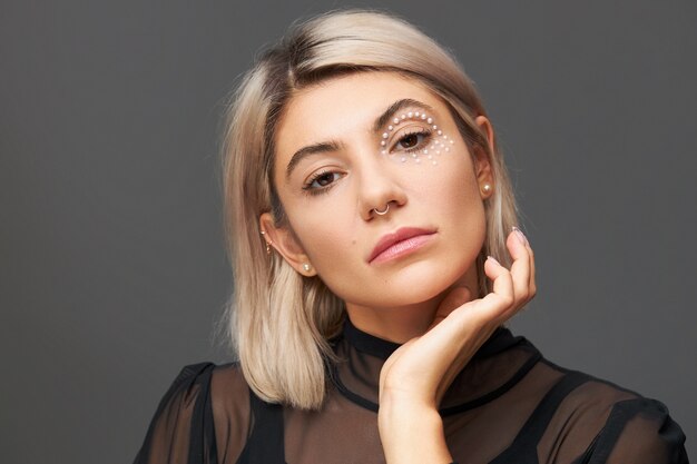 Retrato de hermosa mujer caucásica joven con elegante corte de pelo, anillo en la nariz y cristales blancos alrededor de un ojo manteniendo la mano en la cara. Cuidado de la piel, maquillaje, cosmética, cosmetología y concepto de belleza.