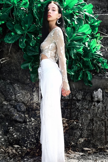 Retrato de hermosa mujer caucásica modelo con cabello largo y oscuro en pantalones clásicos de pierna ancha posando cerca de rocas y fondo de hojas exóticas tropicales verdes