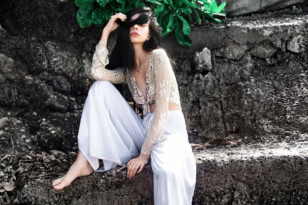 Retrato de hermosa mujer caucásica modelo con cabello largo y oscuro en pantalones clásicos de pierna ancha posando cerca de rocas y fondo de hojas exóticas tropicales verdes