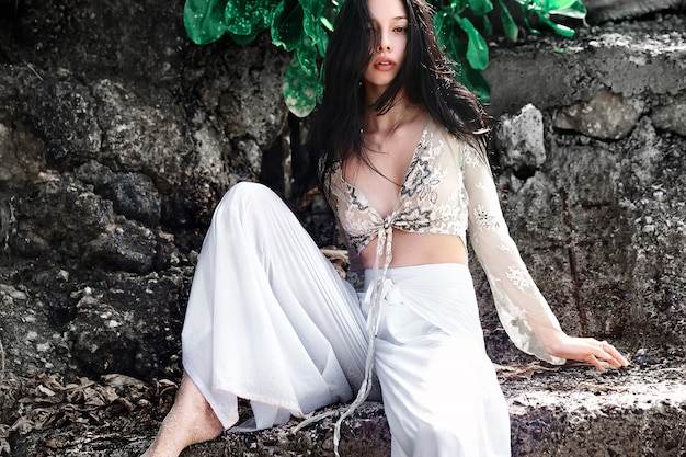 Retrato de hermosa mujer caucásica modelo con cabello largo y oscuro en pantalones clásicos de pierna ancha posando cerca de rocas y fondo de hojas exóticas tropicales verdes