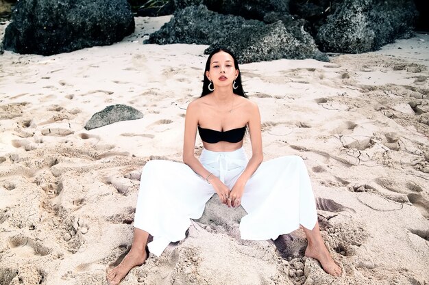 Retrato de hermosa mujer caucásica modelo con cabello largo y oscuro en pantalones clásicos de pierna ancha sentado en la playa de verano con arena blanca cerca de rocas