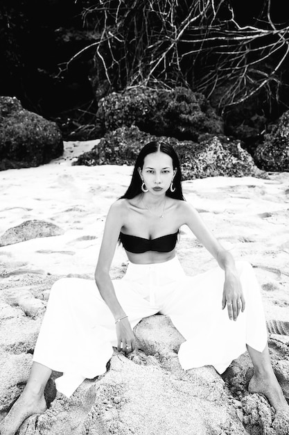 Retrato de hermosa mujer caucásica modelo con cabello largo y oscuro en pantalones clásicos de pierna ancha sentado en la playa de verano con arena blanca cerca de rocas