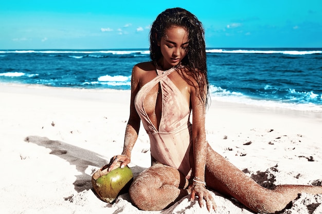 Retrato de hermosa mujer caucásica tomar el sol modelo con cabello largo y oscuro en traje de baño beige posando en la playa de verano con arena blanca en el cielo azul y el océano