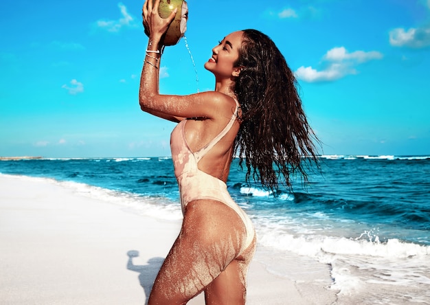 Retrato de hermosa mujer caucásica tomar el sol modelo con cabello largo y oscuro en traje de baño beige posando en la playa de verano con arena blanca en el cielo azul y el océano