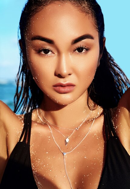 Retrato de hermosa mujer caucásica tomar el sol modelo con cabello largo y oscuro en traje de baño oscuro posando en la playa de verano con arena blanca en el cielo azul y el océano