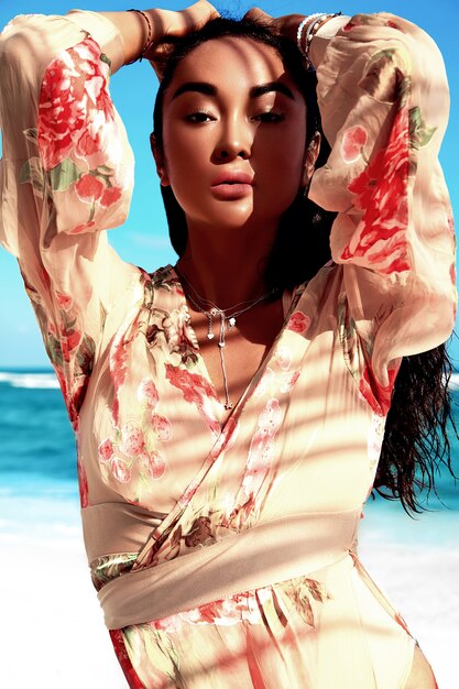 Retrato de hermosa mujer caucásica tomar el sol modelo con cabello largo y oscuro en vestido volador beige posando en la playa de verano con arena blanca en el cielo azul y el océano
