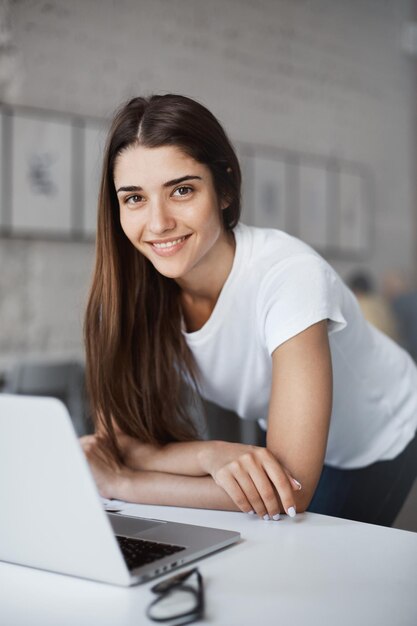 Retrato de una hermosa mujer caucásica usando una computadora portátil parada en un centro de coworking de espacio abierto buscando un nuevo trabajo mirando a la cámara sonriendo