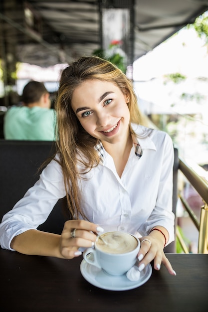 Retrato de hermosa mujer encantadora está revolviendo el café en la cafetería al aire libre y pensando